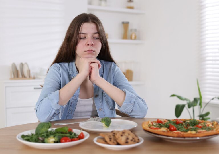 Frau sitzt nachdenklich vor Tisch mit Salat, Pizza und Keksen – Appetitlosigkeit und veränderter Geschmack unter GLP-1-Therapie